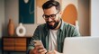 © Mono - Smiling bearded man in glasses using his smartphone while sitting at a desk with a laptop.