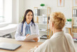 © Studio Romantic - Smiling female doctor in white coat with stethoscope sitting at desk, holding clipboard, talking to elderly patient, providing professional medical consultation in bright organized medical office.