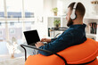 © Prostock-studio - A young man sits in an orange chair, focused on his laptop. He wears headphones and enjoys a bright, airy living room with natural light streaming in.