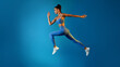 © Prostock-studio - A determined young sportswoman is captured in mid-air, leaping with confidence. She is dressed in a fitness outfit and appears serious as she poses against a vibrant blue background.