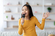 © Prostock-studio - A young girl enjoys singing with a microphone in a bright living room. She smiles, adding joy to a fun afternoon gathering with family or friends.