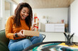 © Prostock-studio - A woman expresses joy and excitement while opening a surprise gift at home. She sits comfortably in a chair, surrounded by a simple and stylish interior.