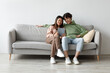 © Prostock-studio - A happy Asian couple sits closely on a gray couch at home, smiling as they engage with a digital tablet. They are exploring a new application together, creating a cozy atmosphere.