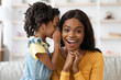 © Prostock-studio - A young girl whispers a secret into her mother's ear while both share joyful expressions. They are in a cozy living room with soft lighting, enjoying a playful moment together.