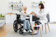 © sofiko14 - A female doctor is giving medication to an elderly woman in a wheelchair, discussing treatment in a home setting.