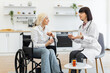 © sofiko14 - A female doctor in a white coat explains medication to an elderly woman in a wheelchair, discussing treatment options in a home setting.