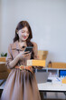 © MINAE - Young Asian woman holding a small package and using a smartphone to manage online orders, a small business, and e-commerce shipping from her home office