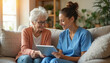 © Vadym - Senior woman smiles looking at tablet with caregiver in blue uniform. Nurse consults elderly patient on couch at home. Medical discussion via device. Home care for old lady.