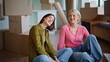 © stockbusters - Tired besties moving house sitting living room laughing between carton boxes