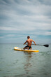 © Aboltin - Man learning to balance on paddle board for the first time. Moment of challenge, focus, and progress under calm sea and cloudy sky.