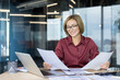 © Liubomir - Experienced businesswoman in glasses at a modern office desk, smiling as she reviews financial documents and reports with laptop and calculator, focused and confident in planning