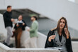 © qunica.com - A professional woman stands at a stairwell talking on her phone, with coworkers blurred in the background. The scene portrays office life, communication, and on-the-go work.
