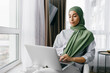 © shurkin_son - Side view of relaxed young beautiful woman in green hijab or headscarf using laptop in her living-room sitting comfortably next to window and watching series or browsing social media news feed