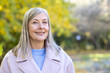 © Liubomir - Senior woman with grey hair smiling confidently at camera in an autumn park, wearing a pink coat and light blue sweater, serene, healthy and enjoying nature's golden foliage