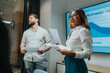 © qunica.com - A confident woman in a white blouse and blue pants leads a presentation while a male colleague holds papers. A large screen displays a chart, highlighting teamwork and a productive office briefing.
