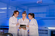 © ultramansk - A team of three young female scientists smiles while working together in a futuristic, blue-lit lab. They are looking at a tablet, sharing ideas and discussing their research.