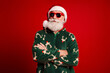 © deagreez - Santa in green christmas sweater with red sunglasses stands confidently against a bold red background
