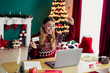 © deagreez - Cheerful woman in red sweater celebrates Christmas at home as she shops online on a laptop beside a twinkling tree and festive decorations