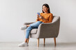© Prostock-studio - A charming young Asian woman sits comfortably in an armchair, using her cellphone to communicate online. She is engaged and focused in a modern setting with a clean background.