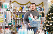 © JackF - Positive young customer standing with shopping basket full of necessary cleaning supplies and toiletries in aisle of cozy decorated household and personal care store during Christmas season