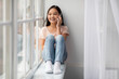 © Prostock-studio - A young girl sits on a windowsill, smiling and chatting on her phone. Sunlight fills the room, creating a cheerful and warm atmosphere. She wears a striped shirt and denim jeans.