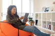 © Prostock-studio - A young woman sits comfortably in an orange bean bag chair, using a tablet in her bright, modern living room filled with books and plants.