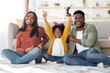 © Prostock-studio - A family sits on a comfortable floor in their cozy living room. The child raises her arms in excitement while holding a gaming controller.
