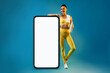 © Prostock-studio - Happy fitness woman in yellow activewear stands by a large smartphone with a blank screen. She holds a water bottle, ready for her workout in a bright blue studio.