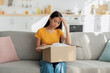 © Prostock-studio - An upset Asian woman sits on a sofa in her home, touching her forehead in disbelief as she looks at an unpacked box. She feels frustrated about receiving the wrong parcel.
