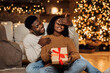 © Prostock-studio - A black man surprises his wife by covering her eyes and presenting a wrapped gift box. They celebrate Christmas and New Year in their beautifully decorated living room.