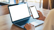© Gravix - A close-up of a person using a laptop and holding a smartphone, both with blank white screens, on a wooden table. Perfect for showing work, mockups, or app use.