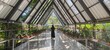 © Creativa Images - Woman standing alone in the center of a modern A-frame greenhouse or plant nursery, looking out at the lush outdoor greenery.
