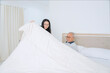 © Creativa Images - Caring Young Woman Tucking Elderly Man into Bed with White Duvet at Home