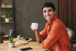 © ViDi Studio - Young man he wear orange casual clothes hold in hand cup drink tea sits alone at table in coffee shop cafe relax rest in restaurant during free time indoors. Freelance mobile office business concept.