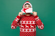 © ViDi Studio - Merry young man wear red Christmas sweater Santa Claus hat posing hold gift coupon voucher card for store show thumb up isolated on plain green background Happy New Year celebration holiday concept