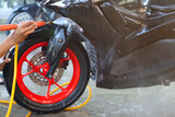 A person using a high-pressure nozzle to spray water on a scooter's black tire and vibrant red alloy rim, cleaning the front wheel during a wash.