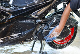 Person washing a glossy black scooter body and rear wheel, covered in thick white soap suds, highlighting vehicle maintenance and care process.
