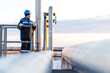 © Кирилл Горшков - Male worker in blue helmet and jacket stands on platform, overseeing industrial pipeline system, with pipes extending into the distance under a clear sky, showcasing safety and vigilance. copy space