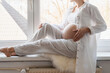 © nataliaderiabina - Side view of beautiful pregnant woman holds hands on belly by the window at home. Pregnancy, maternity, preparation and expectation concept. Beautiful tender mood photo of pregnancy.