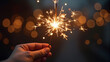 © Andry - A person's hand holding a brightly burning sparkler, creating a festive atmosphere with warm, glowing bokeh lights in the background