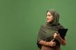 © Prostock-studio - A woman in a hijab stands in front of a dark green background, holding a clipboard while looking to the side. She appears focused and is likely preparing important documents or reports.