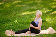 © GRON777 - Middle-aged woman meditates while sitting on fitness mat in grass outdoors. Woman performs hip stretches, practicing mindful movements during outdoor yoga with emphasis on breathing, correct posture.