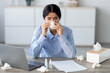 © Prostock-studio - A young Hindu female manager is sitting at her home office desk. She blows her nose in a napkin, showing discomfort from a headache and cold while trying to work online.