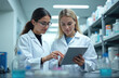 © Maryna - Two women scientists review experiment results on tablet in lab. One points at screen showing data, discussing findings with colleague. Shelves with medical supplies in background. Teamwork in
