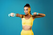 © Prostock-studio - A determined fitness woman is exercising with dumbbells while wearing yellow gym attire. She is flexing her arm muscles and is set against a vibrant blue background, showcasing her strength.