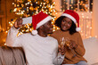 © Prostock-studio - A young African American family enjoys a festive moment together on the sofa. Wearing Santa hats, they cheer with champagne glasses while taking a selfie in front of a decorated Christmas tree.