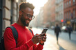 © Maryna - Smiling african american man uses smartphone typing message standing outdoors. Cheerful black guy wearing red sweater looks at phone screen in city street. Happy male model enjoys mobile tech.