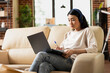 © DC Studio - Professional asian woman sits on couch with notepad and laptop, reviewing notes for business strategy. Focused female individual working from home as virtual assistant, writing checklist for employer.