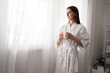 © supAVADEE BUTRADEE - A beautiful young woman, wearing a bathrobe, wakes up early, relaxes, drinks a refreshing coffee, and stands by the window in the morning.