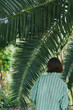 © SHOTPRIME STUDIO - Woman in a striped green shirt walks beneath lush palm leaves, enjoying a serene tropical setting. Soft light filters through fronds, creating calm green and gold tones around the figure.